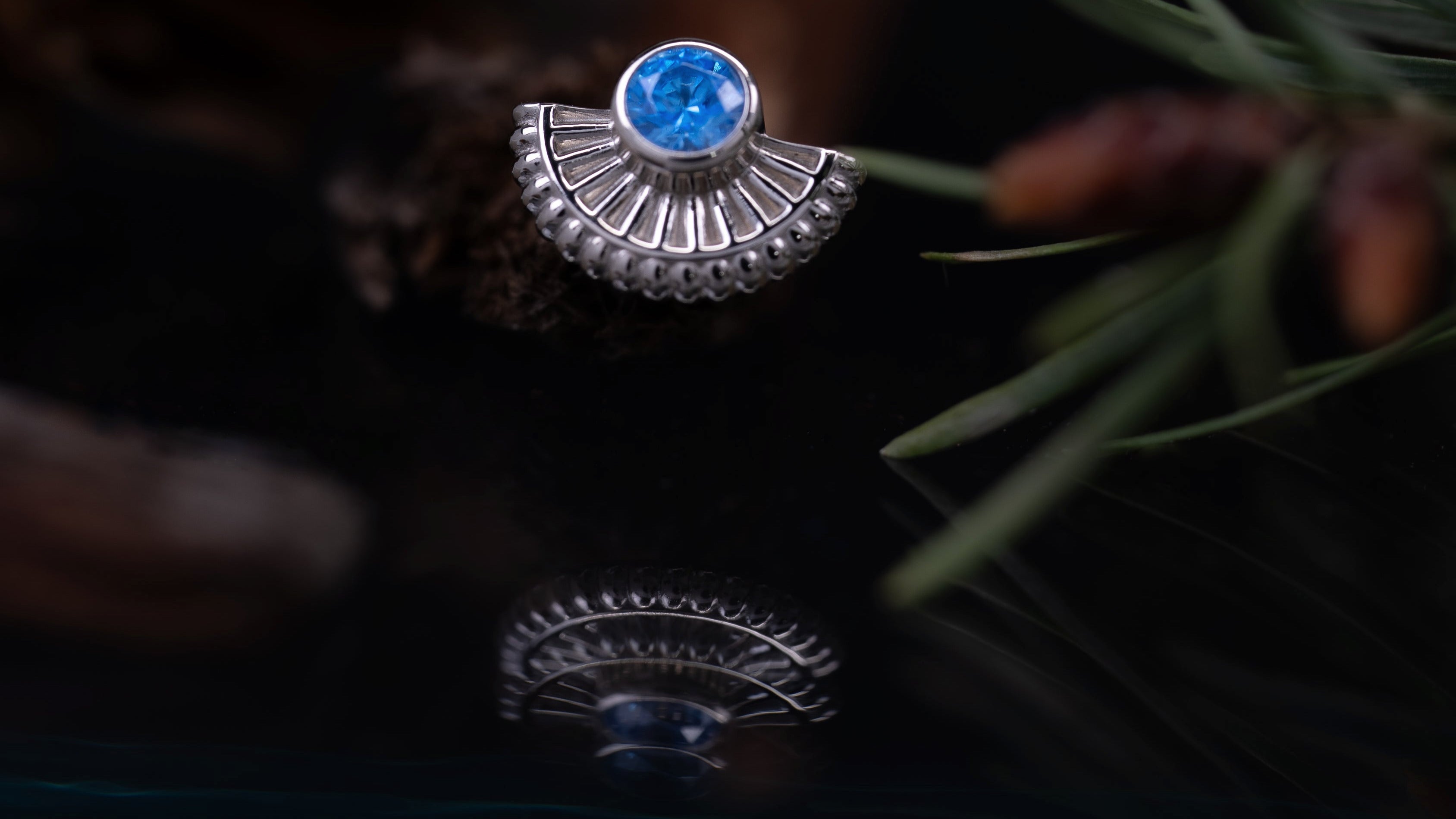 Silver ring with a blue gemstone on a dark background with pine cones and branches.