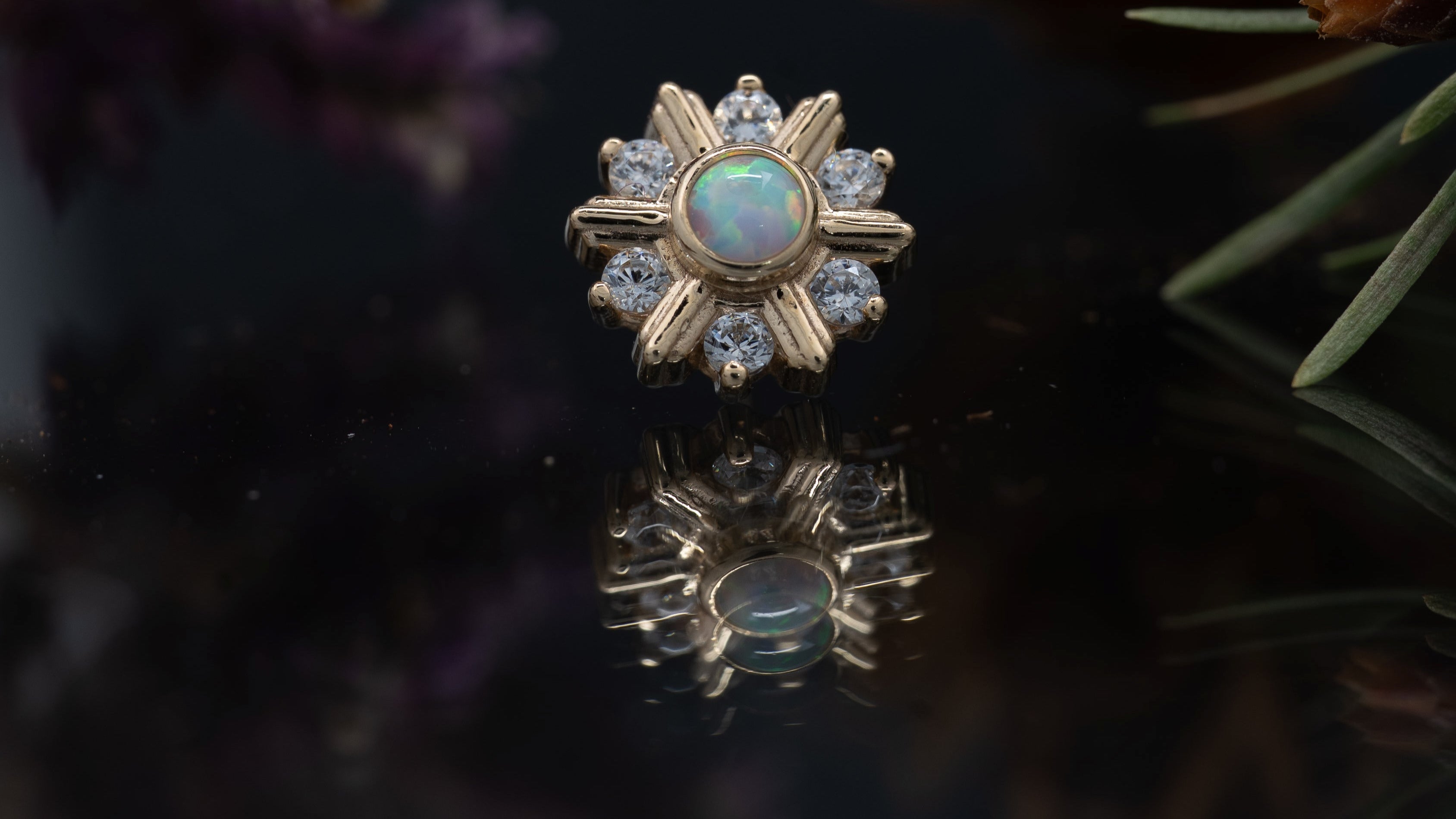 Gold ring with a sunflower design and opal stone on a reflective surface with flowers in the background