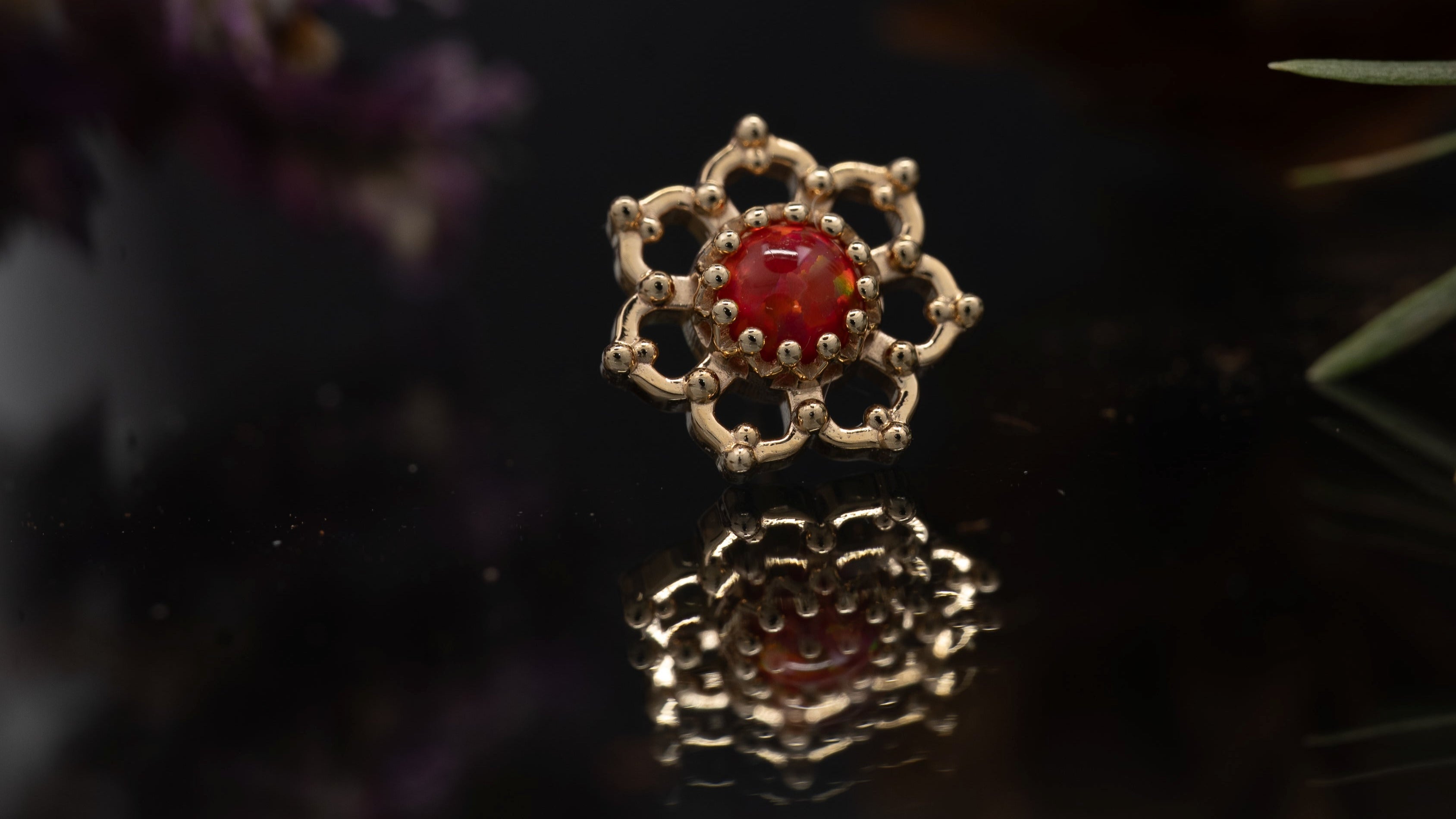 Gold earring with a red gemstone on a reflective surface with flowers in the background